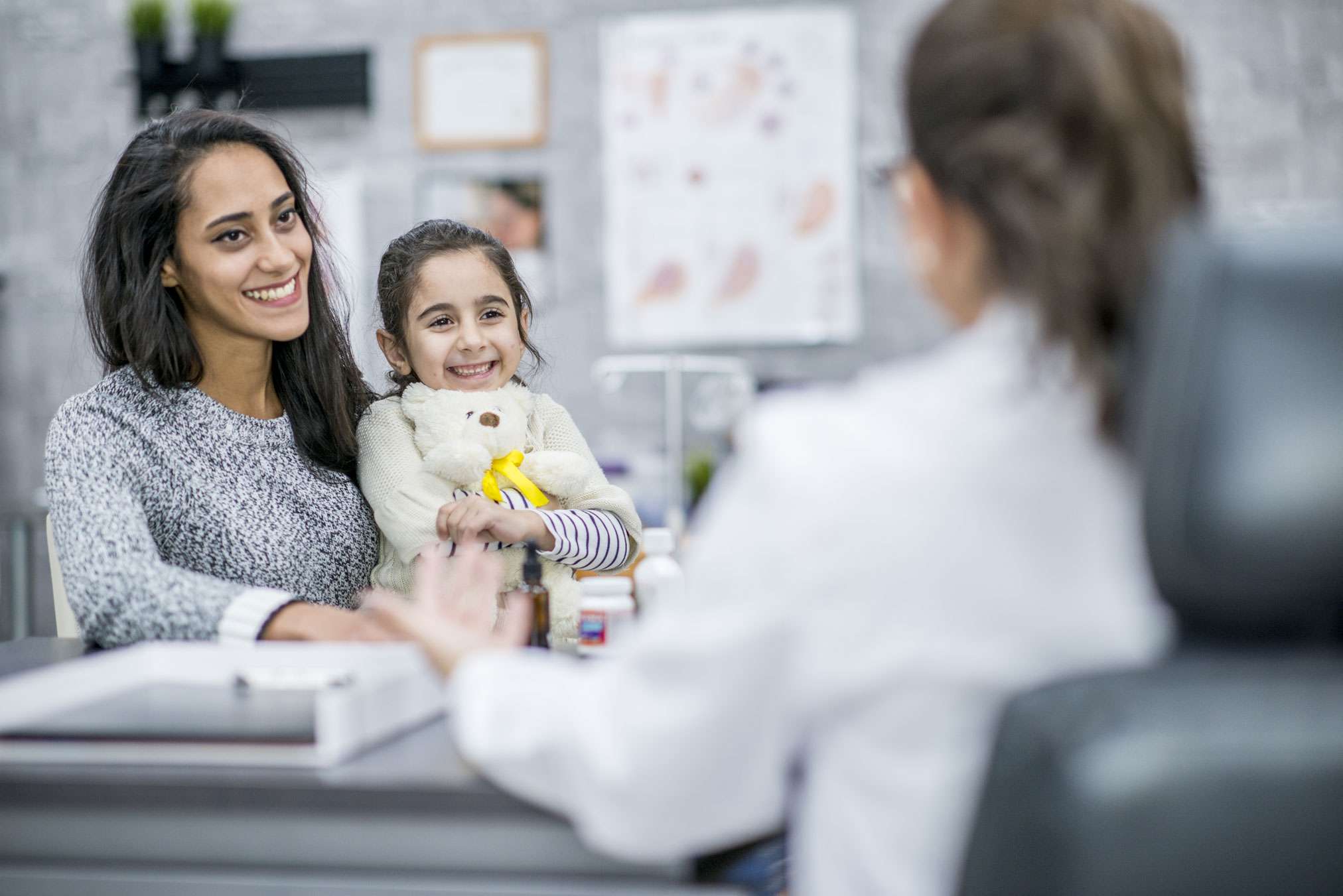 Photo of woman holding a small child and talking to a doctor in the doctor's office. The child is smiling and holding a teddy bear
