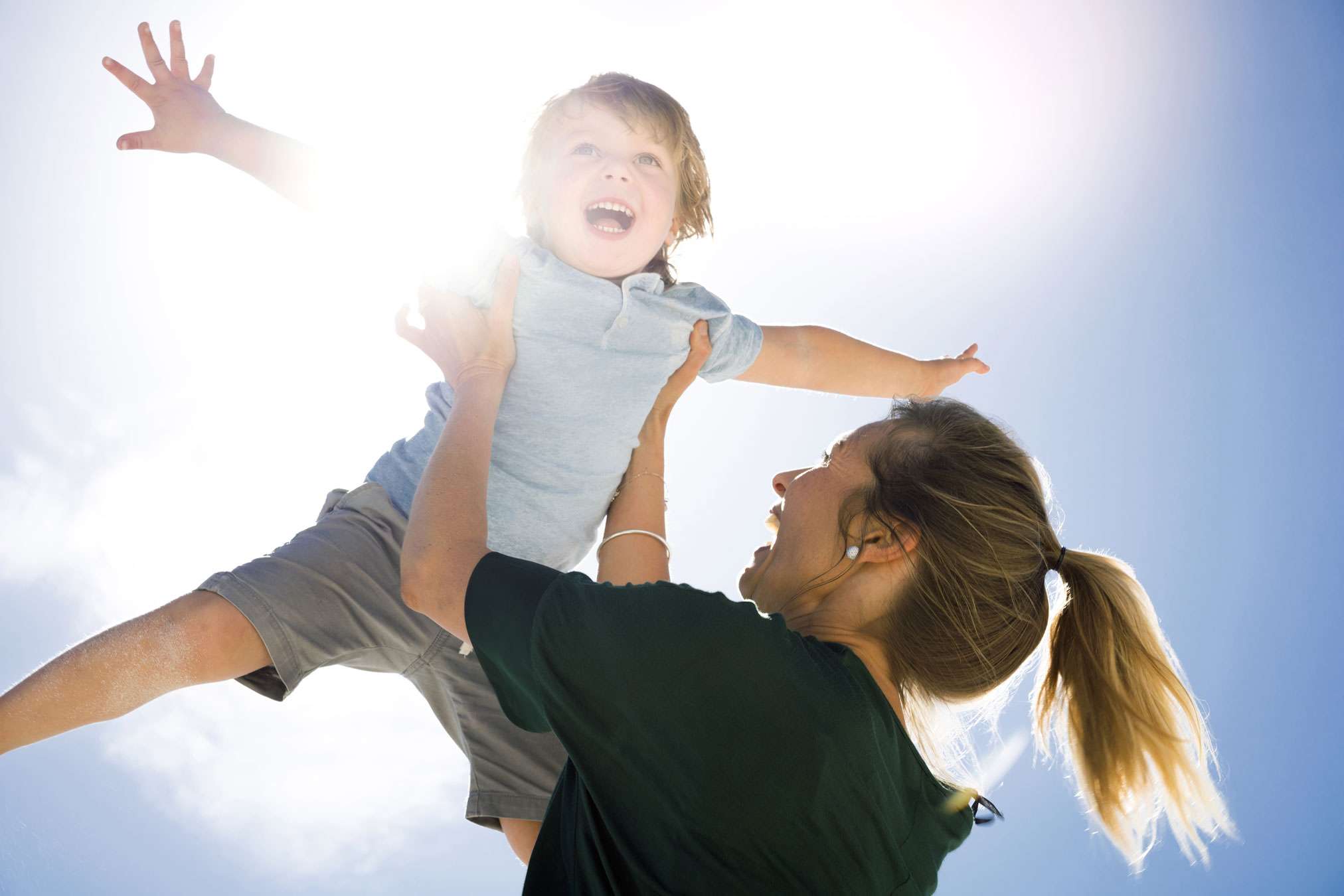 Color photo of woman holding up a 5 year old laughing boy against a blue sunny sky. He is wearing a light blue shirt and gray pants and has he arms out stretched as if he's flying. She is wearing a dark t-shirt and has a blonde ponytail. She is laughing.